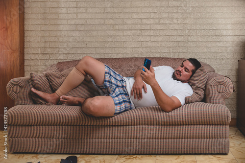 An unemployed and lazy man watching videos, playing mobile games or on social media on his cellphone all day while lying on the couch recovering from a calf injury. photo