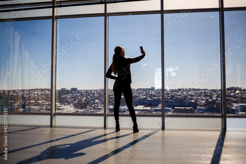 A beautiful girl takes a selfie on her phone against the background of panoramic windows in a skyscraper