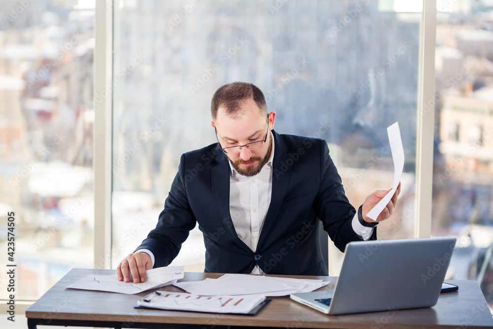 Adult successful male businessman, teacher, mentor is working on a new project. Sits at the table by the large window. It works on a laptop Adult successful male businessman, teacher, mentor is working on a new project. Sits at the table by the large window. It works on a laptop