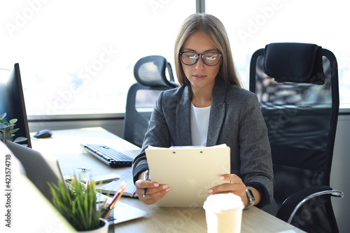 Attractive business woman holding documents and looking at them while sitting at the desk in office