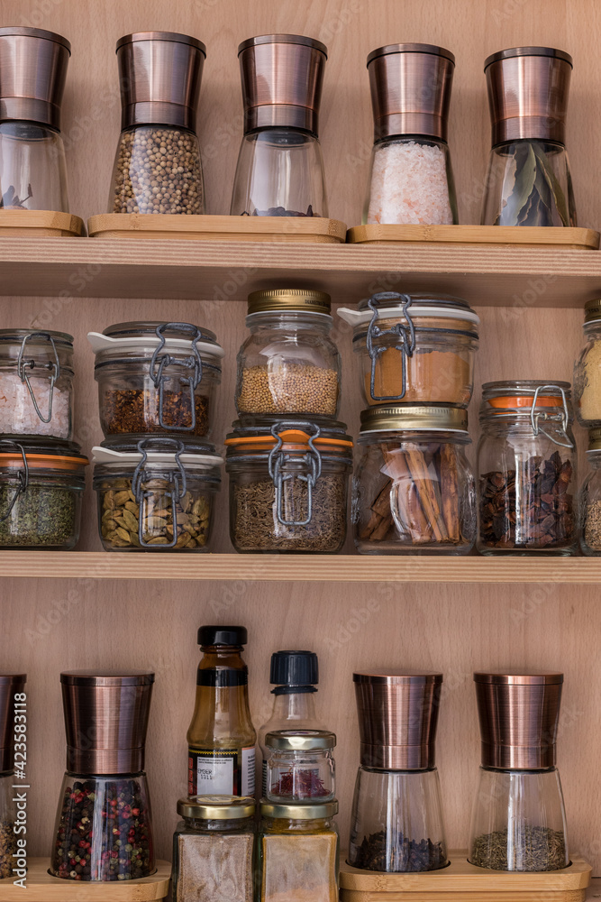 Glass piled jars and manual copper elegant mills full of spices, salt, aromatic plants over wooden kitchen selves.