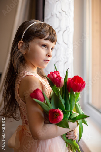 A Caucasian six-year-old girl in a pink dress holds red tulips in her hands and stands at the window