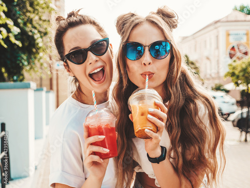 Two young beautiful smiling hipster female in trendy summer clothes. Carefree women posing outdoors.Positive models holding and drinking fresh cocktail smoothie drink in plastic cup with straw photo