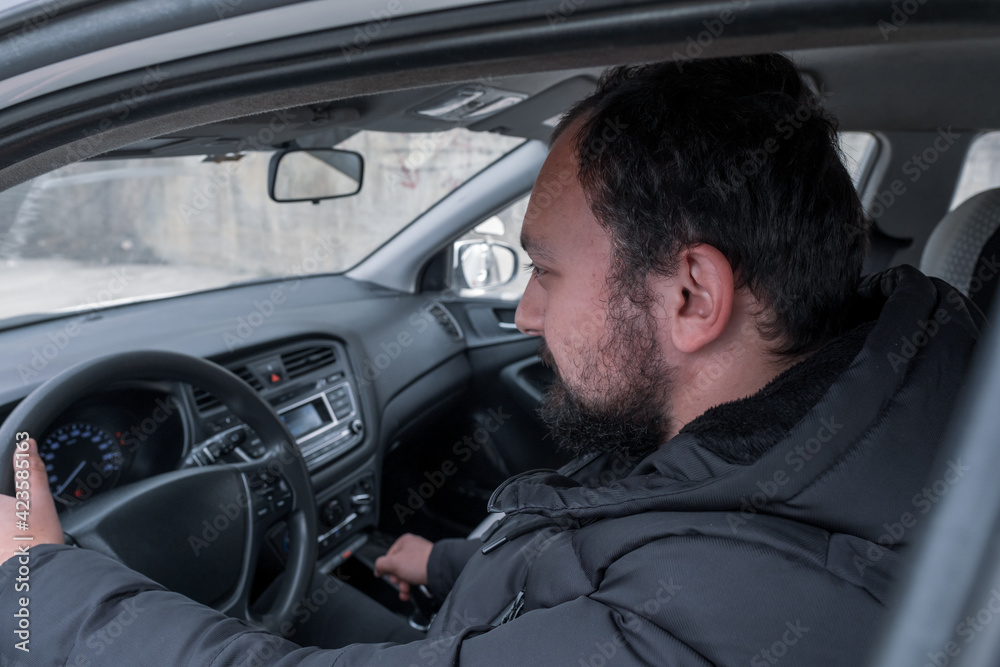 one black bearded young man holds gear shift and drives a car and looks at road in daylight