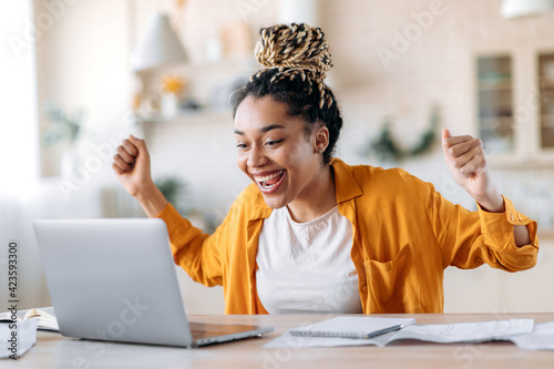 Overjoyed excited african american girl with dreadlocks, freelancer, manager working remotely at home using laptop, looks at screen with surprise, smiling face, gesturing with hands, got a dream job photo