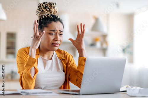 Tense indignant frustrated african american girl with dreadlocks, business woman, manager or freelancer working remotely at a laptop, experiencing stress at work, got bad message, gesturing with hands
