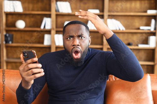 Shocked black man by the bad news he saw on the mobile phone, looking at camera, businessman sitting on the couch with open mouth, gesturing with hand, isolated over the background of bookshelves photo