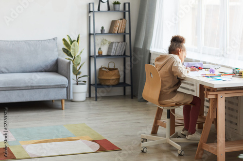 Full length portrait of cute African-American girl doing homework or drawing while sitting at desk in cozy home interior, copy space