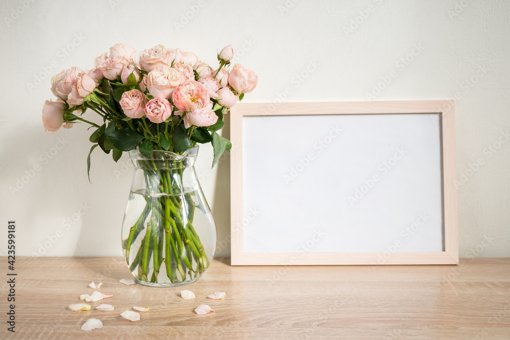 Portrait white picture frame mockup on wooden table. Modern glass vase with roses. White wall background. Scandinavian interior. 
