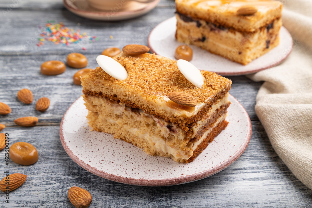 Honey cake with milk cream, caramel, almonds and a cup of coffee on a gray wooden background. Side view. Honey cake with milk cream, caramel, almonds and a cup of coffee on a gray wooden background. Side view.