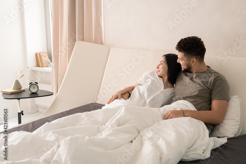 Young affectionate restful couple relaxing on large double bed in the morning and looking through window while chatting about pleasant stuff