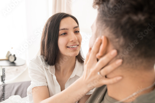 Happy young brunette woman looking at her husband eyes with kind smile while touching his face and wishing him good morning after sleep