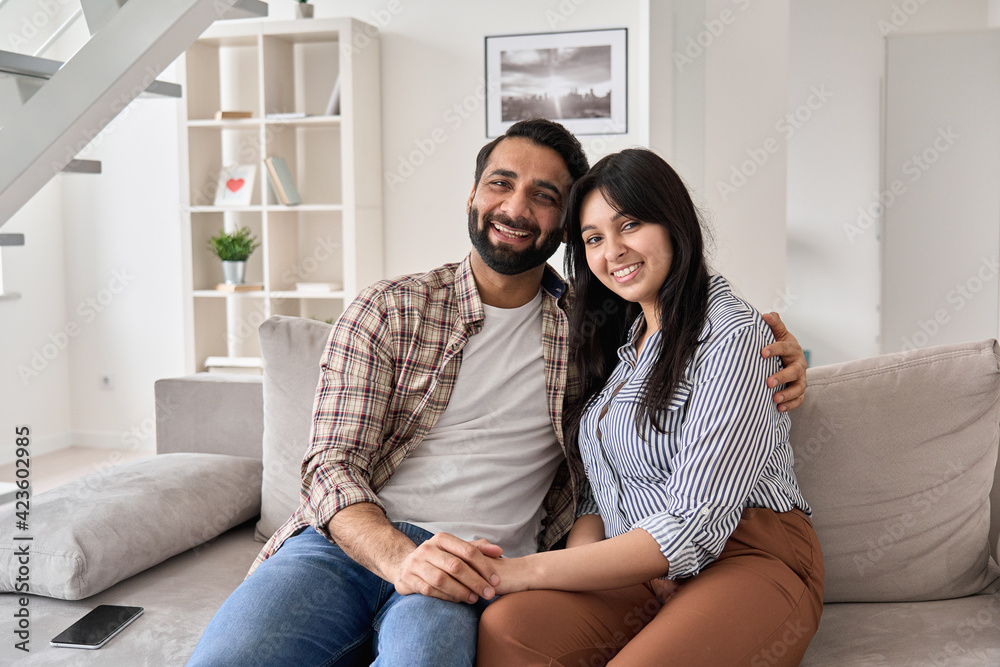 Happy young indian couple real estate buyers hugging sitting on couch at home looking at camera. Smiling husband and wife new homeowners embracing, enjoying own apartment purchase, portrait.