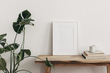 White portrait frame mockups on vintage bench, table. Cup of coffee on pile of books and monstera potted plant. White wall background. Scandinavian interior, neutral color palette. Artistic display.
