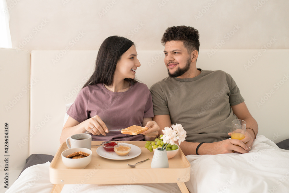 Happy young affectionate couple in t-shirts sitting in bed and talking during breakfast while female spreading peanut butter on toast