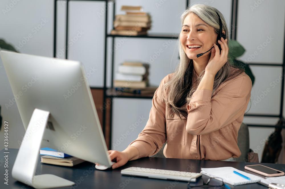 Joyful successful beautiful mature asian business woman call center manager working in office, communicating with business team via virtual conference, video call, using headset, smiling friendly Joyful successful beautiful mature asian business woman call center manager working in office, communicating with business team via virtual conference, video call, using headset, smiling friendly