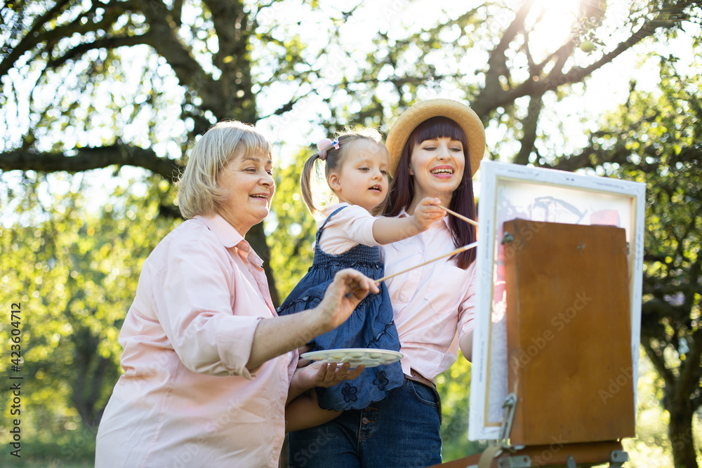Fototapeta premium Easel, canvas, brushes and paints. Family leisure. Close up bottom view of three generation family, grandmother, mother and little girl artist, painting a picture on canvas outdoors together.