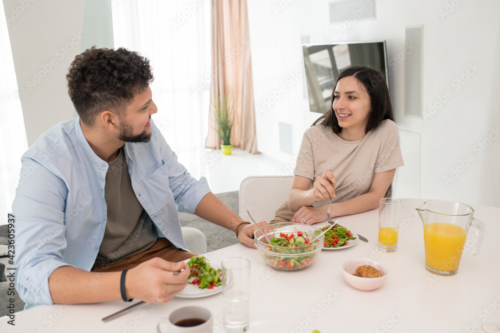 Happy young brunette woman looking at her husband with smile while both having selfmade vegetable salad and orange juice by table