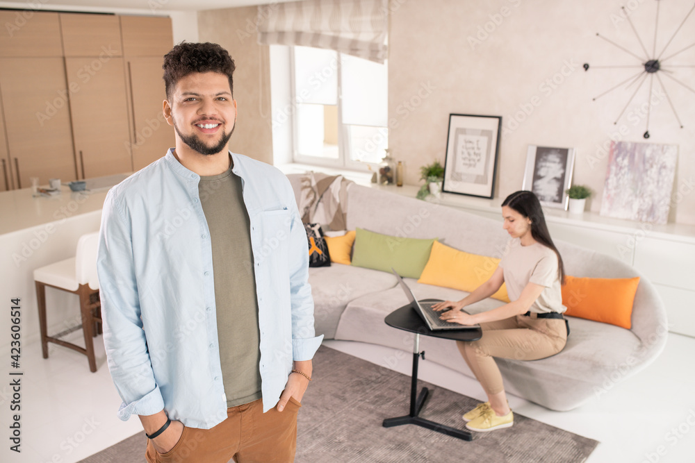 Young cheerful man in casualwear looking at you while standing against his wife sitting on couch and using laptop while networking