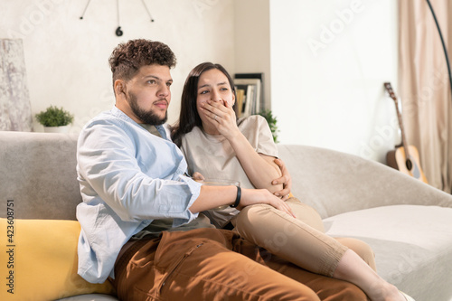 Young scared brunette woman covering mouth by hand while sitting on couch close to her husband and both watching horror movie © pressmaster