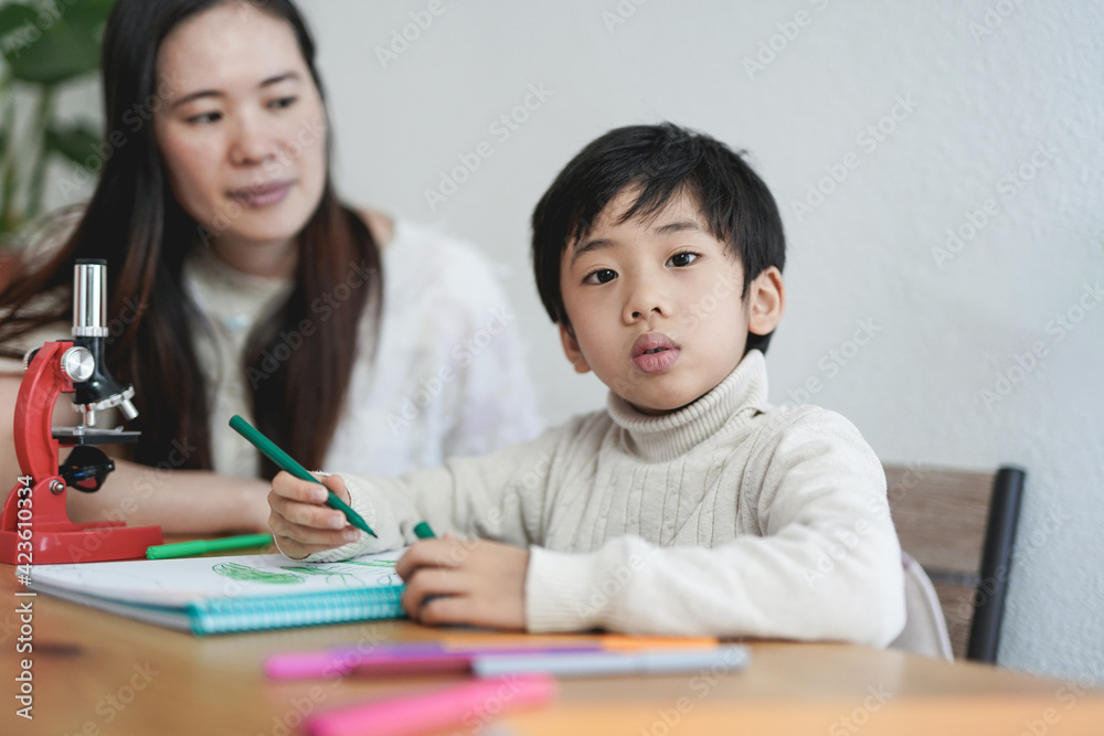 Little asian child looking in camera while doing drawing activities with nanny - Daycare and home schooling Little asian child looking in camera while doing drawing activities with nanny - Daycare and home schooling