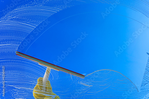 Window cleaner using a squeegee or wiper to wash a window glass. Hand in yellow glove hold cleaning squeegee on a blue sky background. Copy space