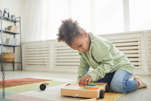 Full length portrait of cute African-American girl playing with toys while sitting on floor at home, copy space