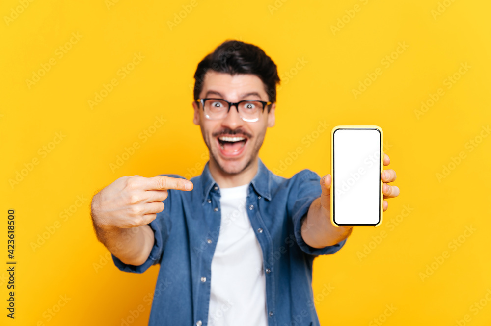 Defocused caucasian unshaven amazed guy with glasses in denim shirt, looks at the camera, holds and points finger at smartphone with blank white screen in hand, copy space, isolated orange background Defocused caucasian unshaven amazed guy with glasses in denim shirt, looks at the camera, holds and points finger at smartphone with blank white screen in hand, copy space, isolated orange background