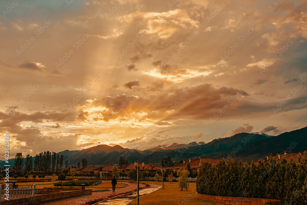 Sunset over resort town with walking man. Mountain landscape is illuminated by rays of sun. Street with identical houses, gardens and lawn. Beautiful clouds. Small fences. Wet sidewalk after rain.