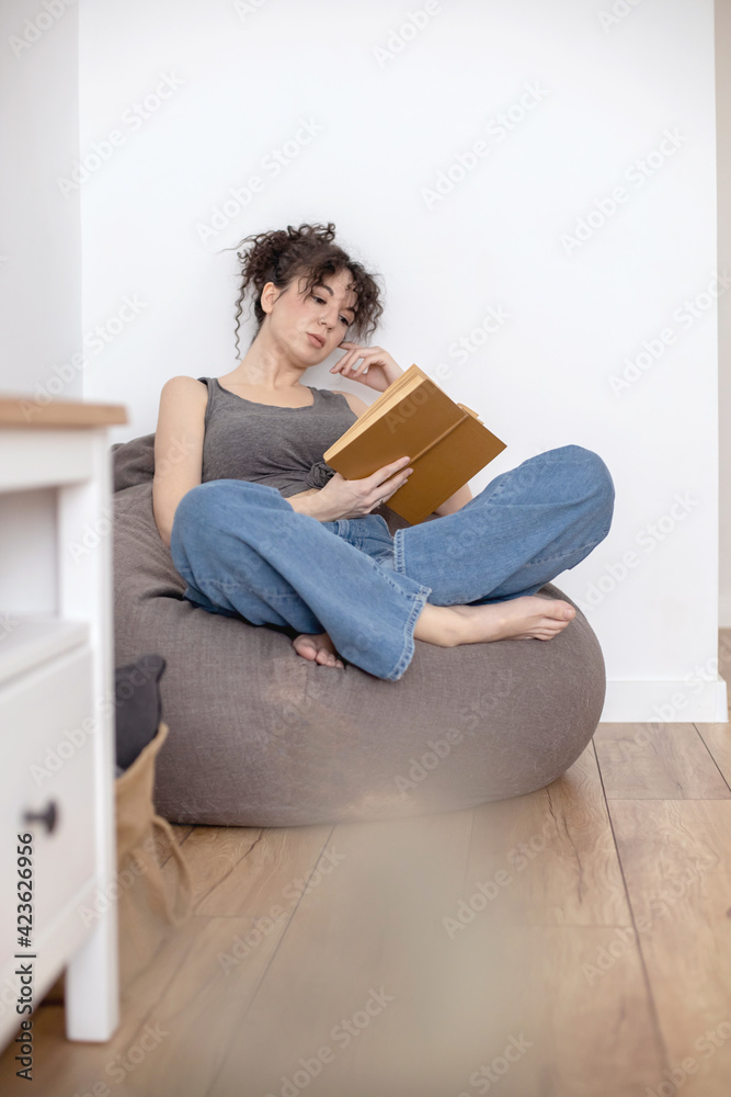 Relaxed brunette woman lying on chair bag reading interesting paper book enjoying weekend at home