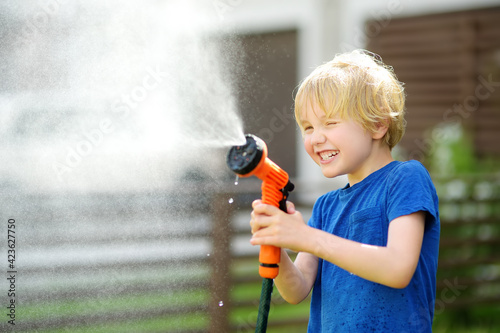 Funny little boy watering lawn and playing with garden hose with sprinkler in sunny backyard. Preschooler child having fun with spray of water.