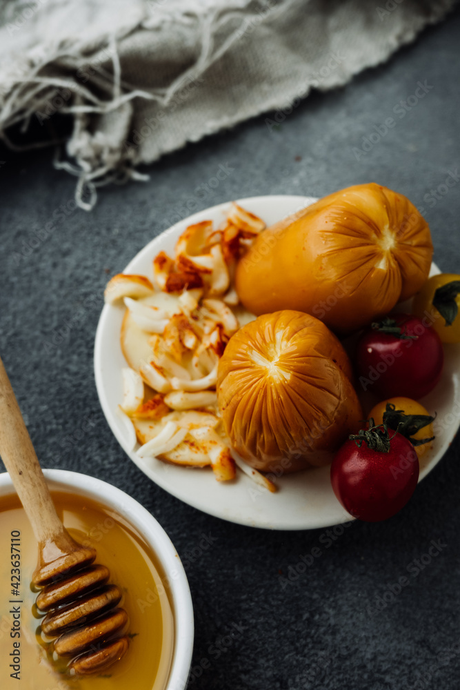 smoked cheese with crust, cheese crumbs and cherry tomatoes on dark table with bowl of honey and wooden stick. types of cheese, options for making products from milk at home, selective focus
