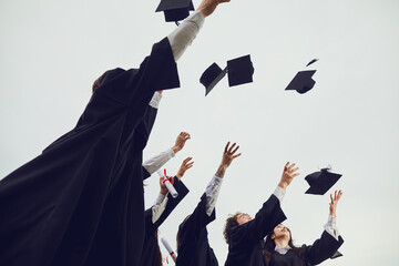 We did it. Group of graduate students in black gowns on sky background throw square academic academic hats at university graduation. Education, graduation and people concept.