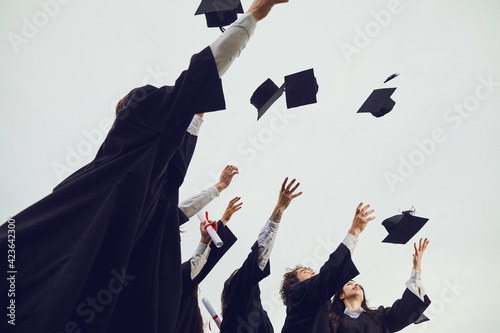 We did it. Group of graduate students in black gowns on sky background throw square academic academic hats at university graduation. Education, graduation and people concept. photo