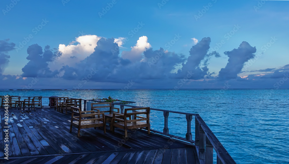 Obraz premium Early morning. On the terrace above the aquamarine ocean, there are tables and chairs damp from the rain. There are picturesque white and blue clouds in the azure sky. Maldivian idyll.