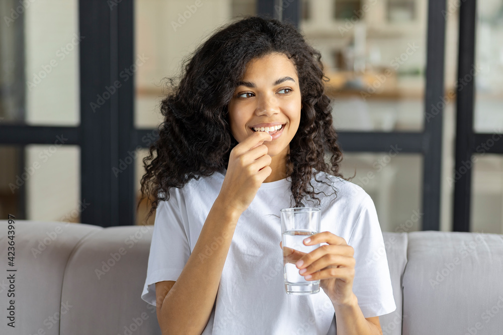 Young beautiful smiling african American woman holding  holding vitamin pill and glass of water sitting  on the couch at home. Healthy lifestyle, healthy diet nutrition concept - obrazy, fototapety, plakaty 
