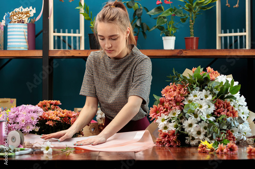 Florist makes a bouquet of chrysanthemums. Young adult girl chooses paper for decoration.