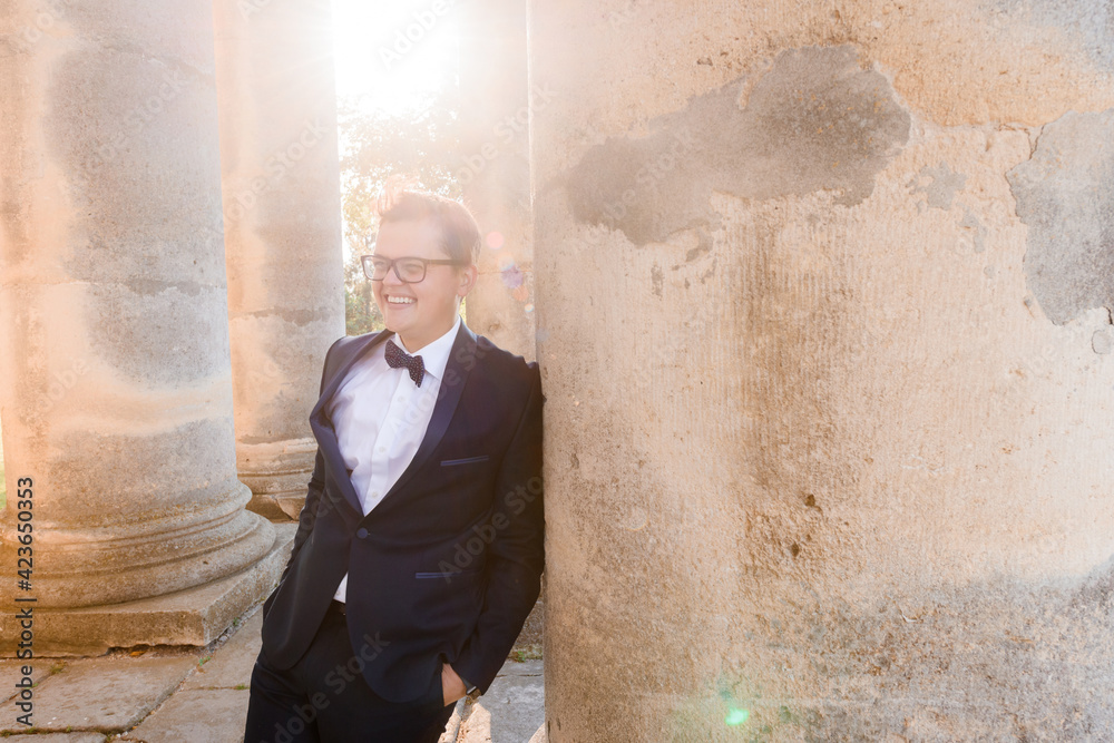 Obraz premium Front view of smiled young male in glasses, wearing in black suit, relying on column of builld, putting hand into pocket of pants and looking away