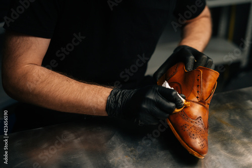 Close-up hands of unrecognizable shoemaker in black gloves rubbing paint on light brown leather shoes with fingers. Concept of cobbler artisan repairing and restoration work in shoe repair shop. © dikushin