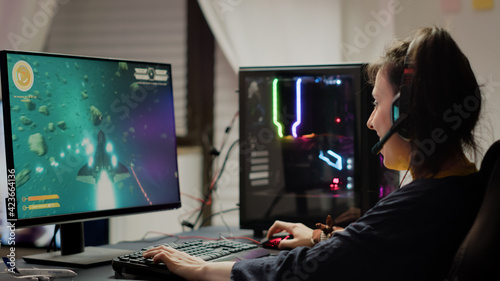 Addicted lady playing space shooter video game using headphones ,wireless joystick and RGB powerful personal computer. Cyber gamer with headset and joypad performing virtual games at gaming