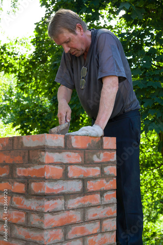 Senior man is making a chimney of red bricks, working with a trowel and making masonry works on a roof of a house, vertical
