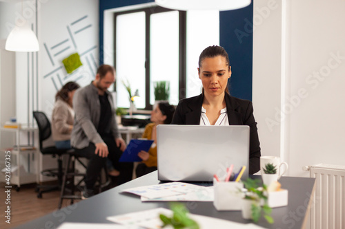 Happy hispanic lady typing on laptop sitting at desk in start up business office drinking coffee while diverse colleagues working in background. Multiethnic coworkers planning new financial project.