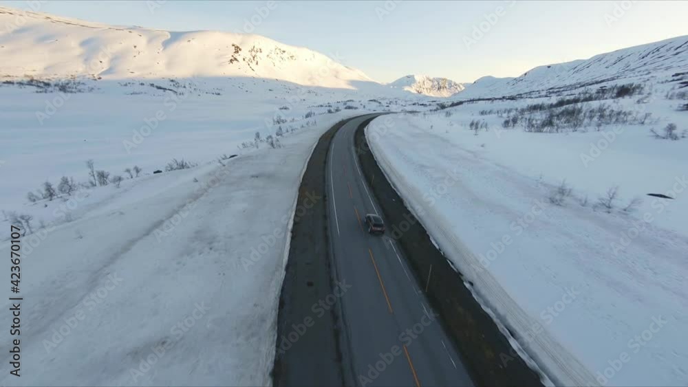 Racing fpv drone following chasing a fast car along a snow coverd mountain road into the sunset, rock and mountains in the background and a frozen lake blue sky aerial Racing fpv drone following chasing a fast car along a snow coverd mountain road into the sunset, rock and mountains in the background and a frozen lake blue sky aerial