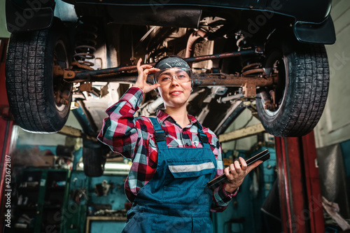 A young pretty smiling female mechanic, in a uniform and glasses, with a tablet in her hands, poses standing under a car on a lift. Indoors garage