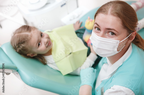 Female dentist in medical face mask looking to the camera while working with a child