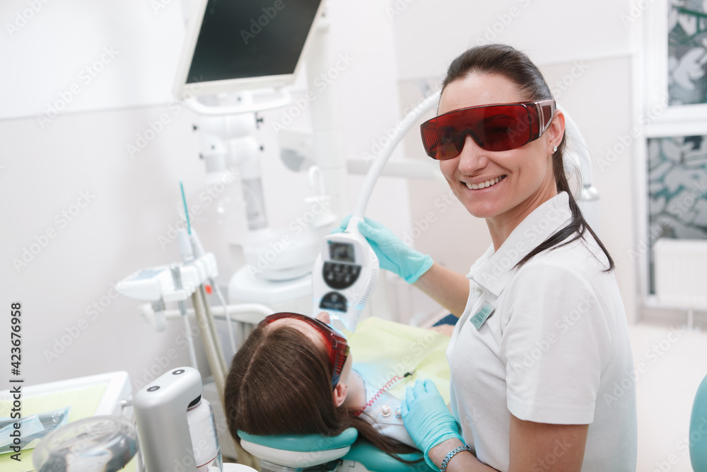 Cheerful female dentist smiling to the camera while whitening teeth of a patient, copy space Cheerful female dentist smiling to the camera while whitening teeth of a patient, copy space