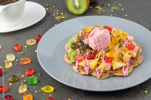 Heart waffle with kiwi and pineapple with gummy candy and ice cream on it.