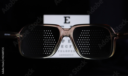 Punched eyeglasses for vision training. Glasses for vision correction. Medical pinhole glasses. Perforated training glasses, eye exam chart on the background, bad eyes concept photo