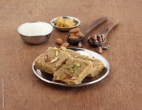 Rava burfi, a delicious indian sweet, made from ingredients like rava or semolina, jaggery, almonds, and pistachios, on a white ceramic plate on a wooden background.