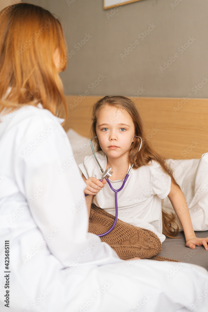 Adorable sick little girl playing with stethoscope while sitting in bed in bedroom during visit of pediatrician at medical clinic, hospital, home. Concept of children health. Adorable sick little girl playing with stethoscope while sitting in bed in bedroom during visit of pediatrician at medical clinic, hospital, home. Concept of children health.